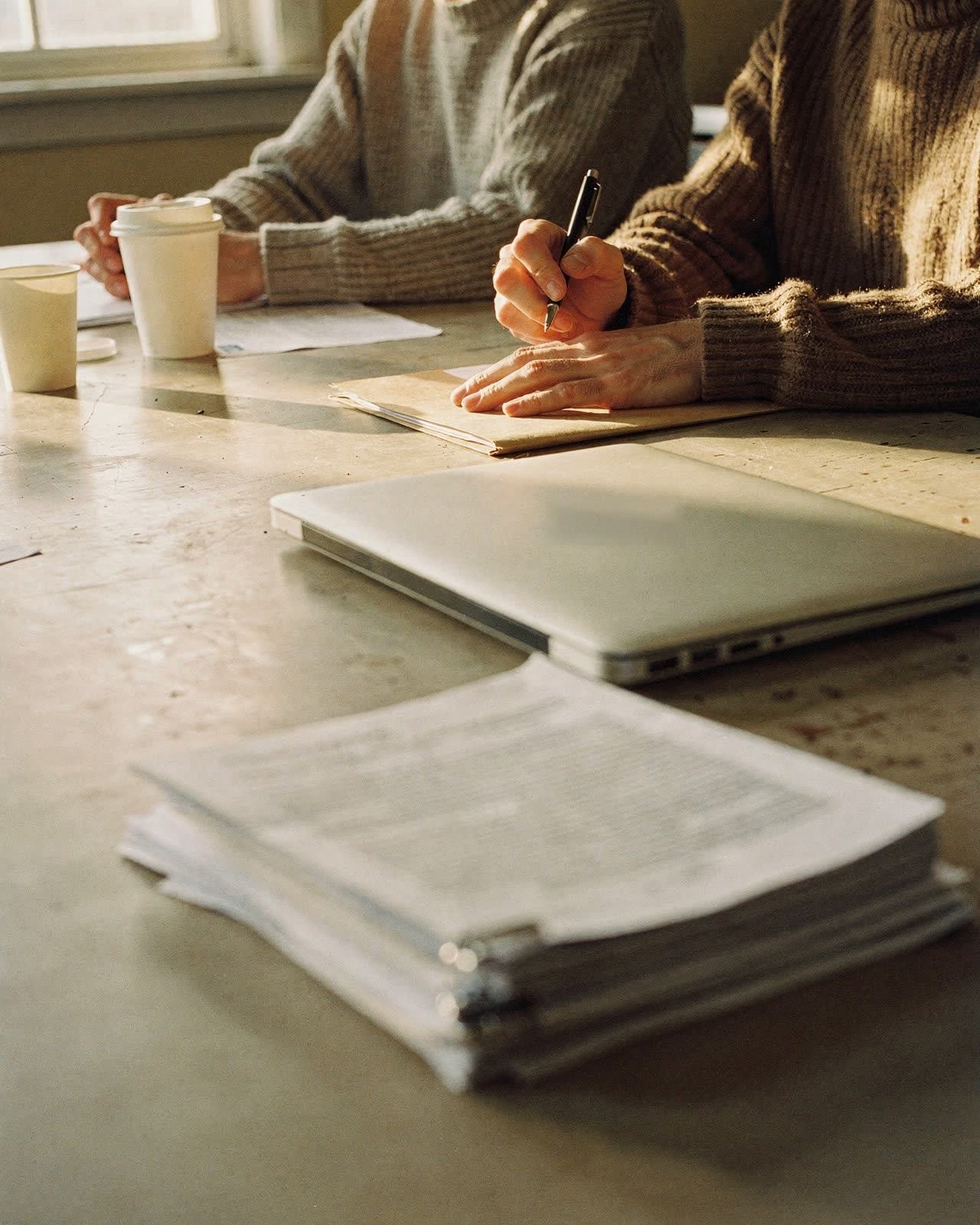 People at a meeting table with coffee, a folder and a stack of printed reports, one taking notes